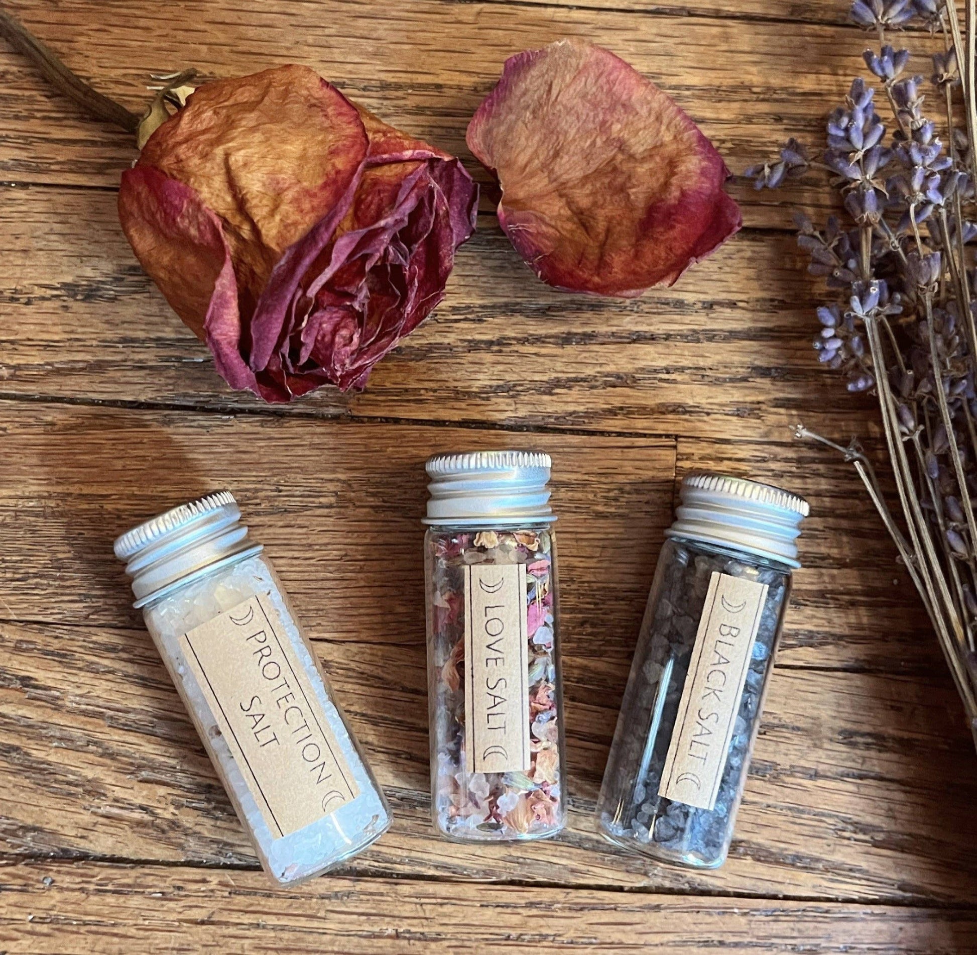 Three small containers labeled protection, love, and black salt containing ritual salt and herbs on a wooden surface with dried flowers.