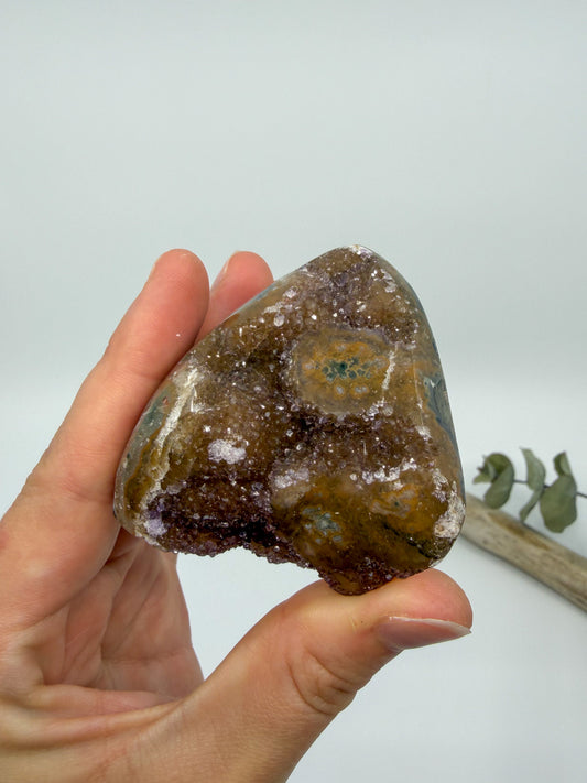 A hand holding a polished rainbow amethyst crystal geode specimen with lavender brown crystalline formations and hints of blue-green inclusions, set against a plain light gray background with a small eucalyptus branch in the corner.