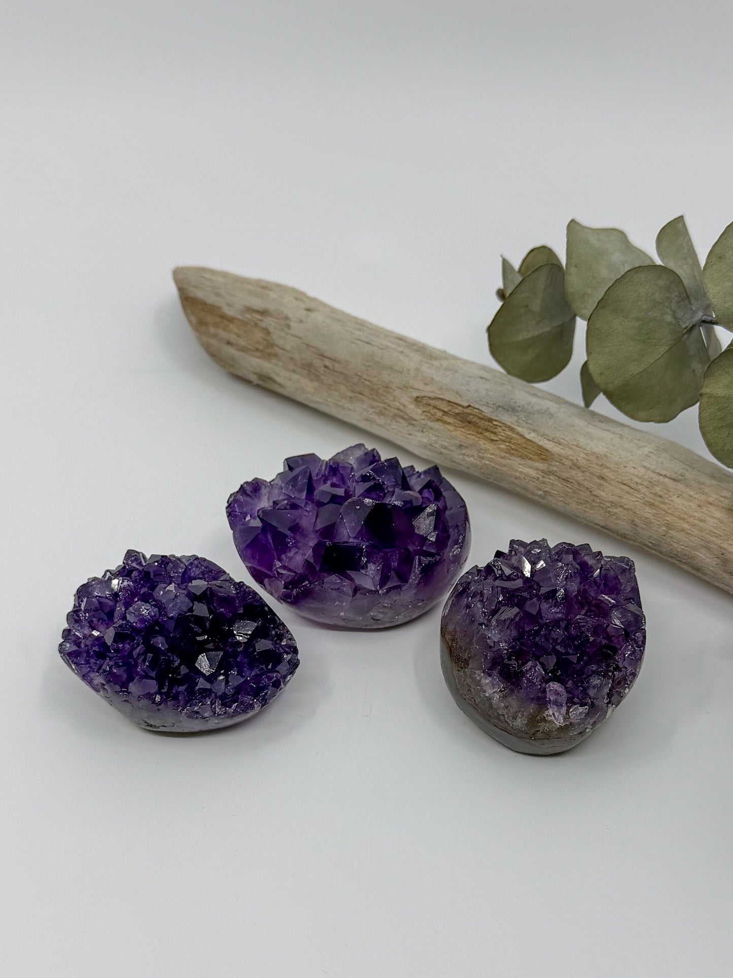 Three polished amethyst crystal geode clusters in shades of purple and violet, arranged on a white surface with a driftwood branch and eucalyptus leaves in the background.