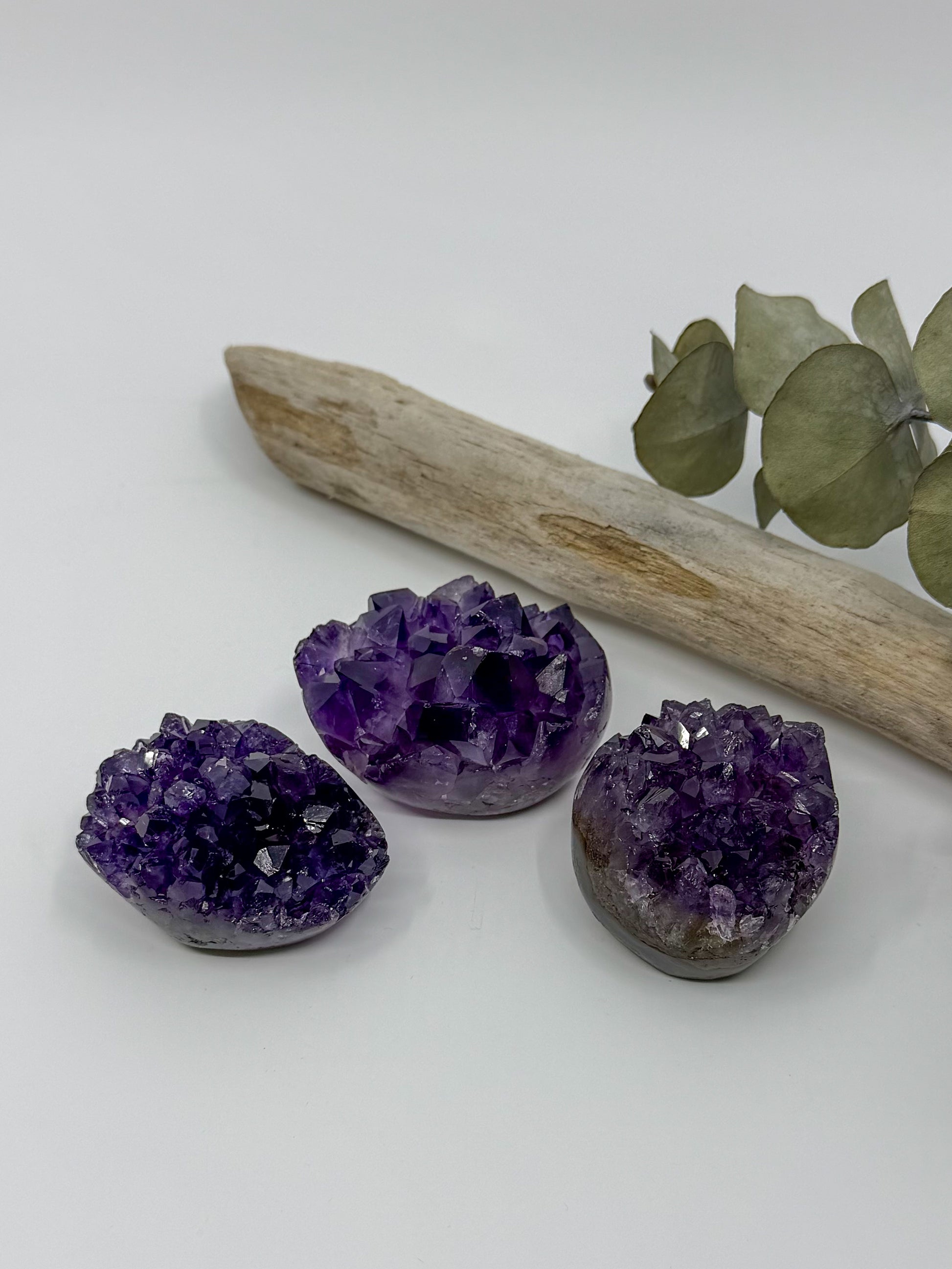Three polished amethyst crystal geode clusters in shades of purple and violet, arranged on a white surface with a driftwood branch and eucalyptus leaves in the background.