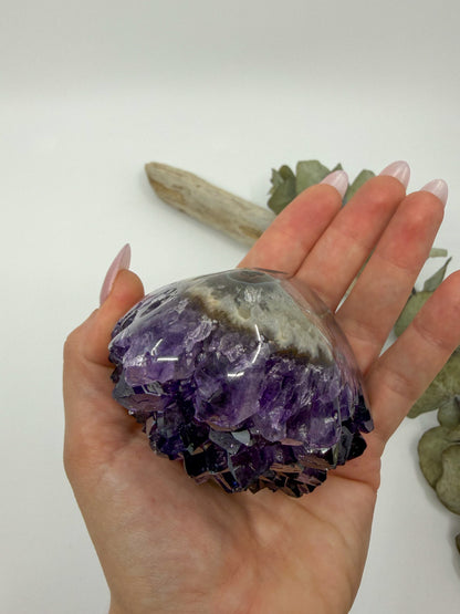 A hand holding a large purple amethyst crystal cluster with a gray and white band near the top, against a plain light background with a piece of driftwood and dried leaves in the distance.