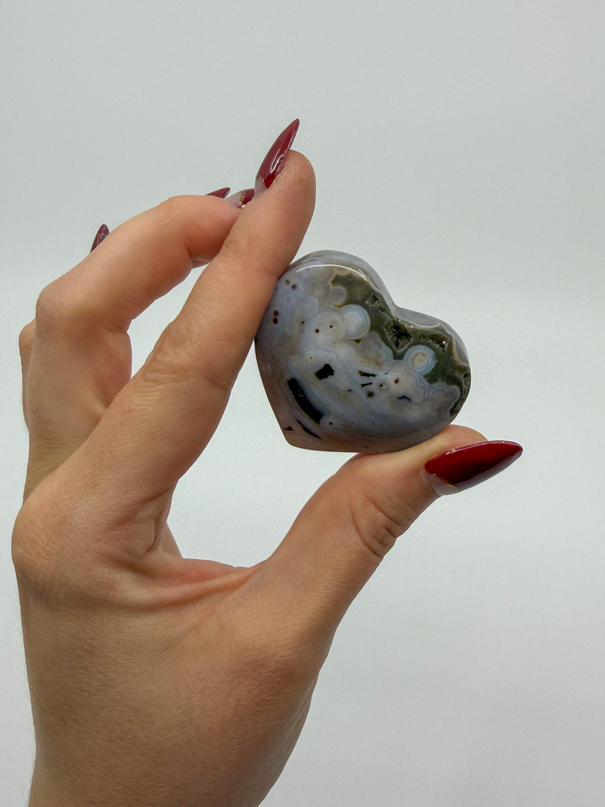 A hand with red manicured nails holds a polished, heart-shaped ocean jasper stone featuring natural marbled patterns and orbs of gray, white, and dark green against a plain light gray background.