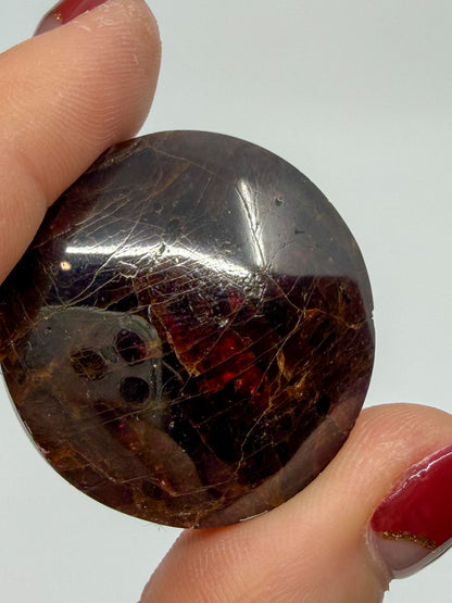 A close-up of a hand holding a polished, oval-shaped dark reddish brown garnet gemstone with visible natural inclusions and a glossy finish against a plain light background.