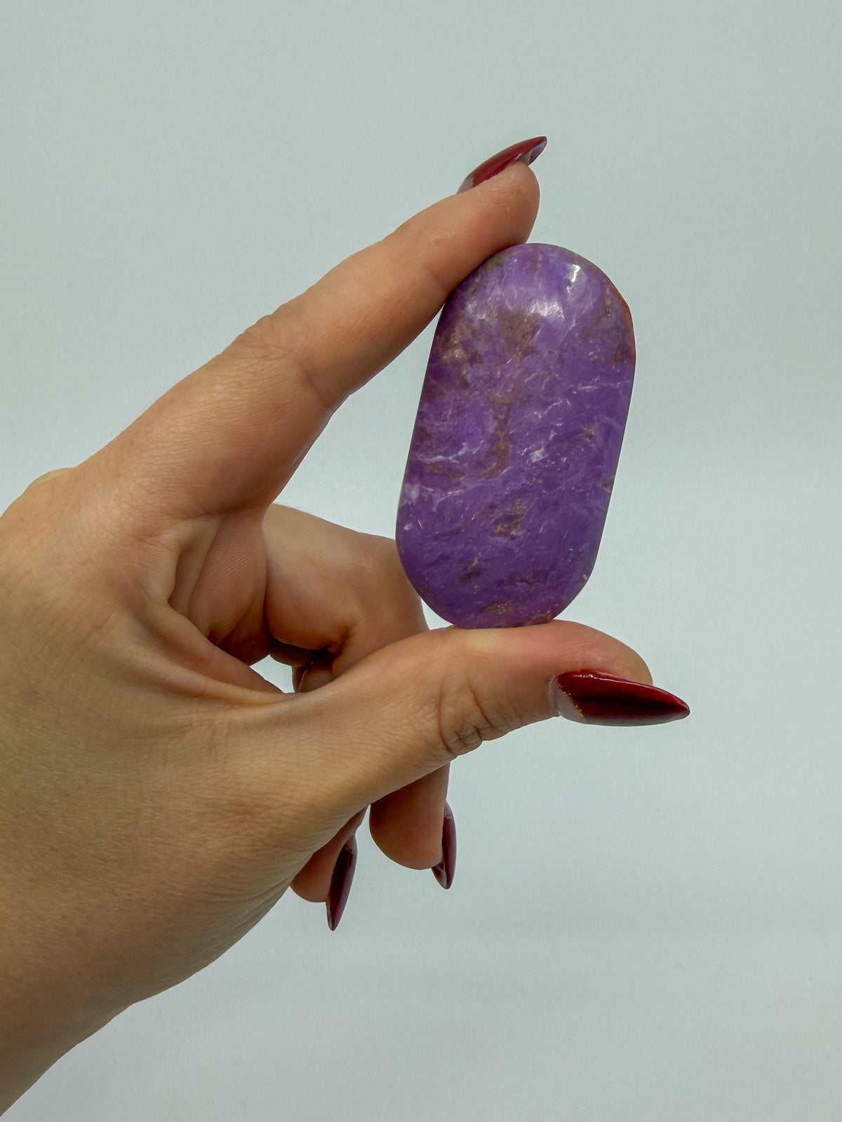 A hand with dark red manicured nails holds a polished purple phosphosidierite stone with natural brown veining against a plain light gray background.